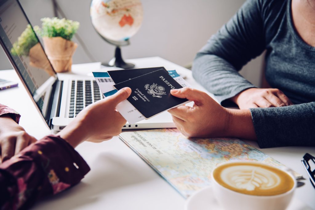 Uma pessoa entregando um passaporte sobre uma mesa com um café, um computador e um mapa