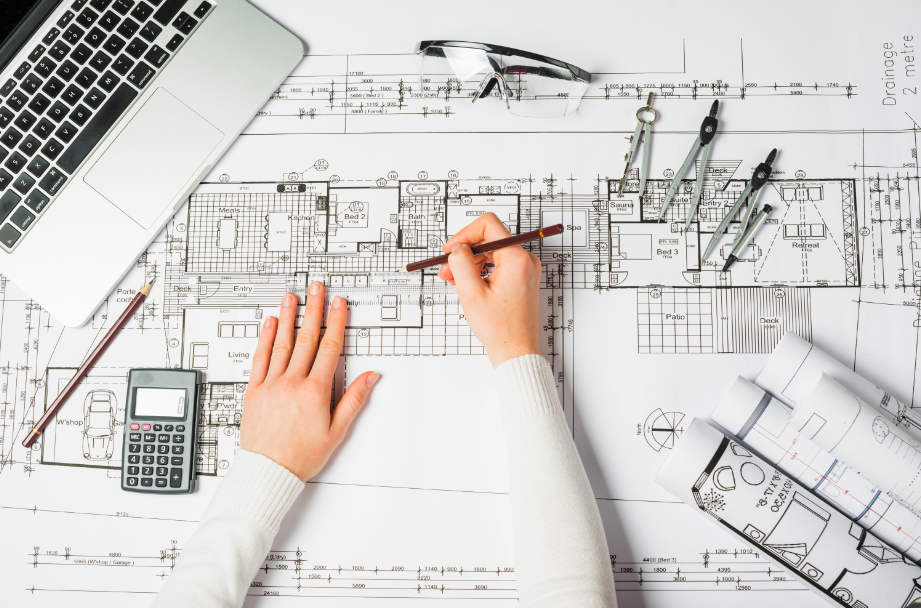 Mão sobre um projeto de arquitetura com um computador, uma calculadora e compassos. Estudar arquitetura no exterior.