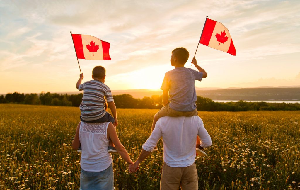 Casal de mãos dadas enquanto os filhos estão em seus ombros balançando bandeiras do Canadá ao pôr do sol. Family day.