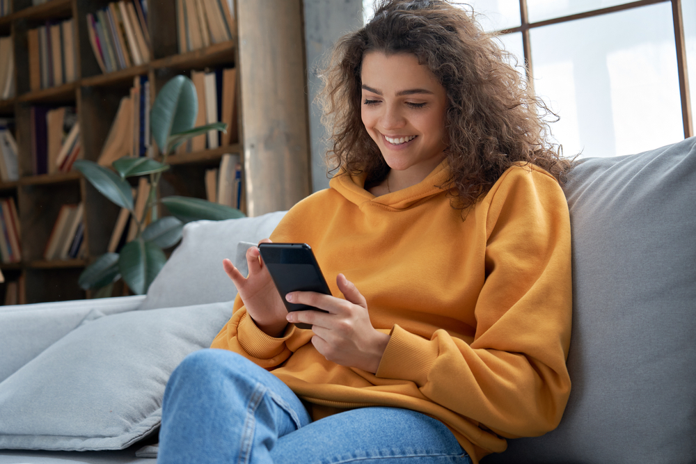 Mulher sentada no sofá com expressão alegre ao utilizar o celular. Como funciona o Sucesso do Estudante.